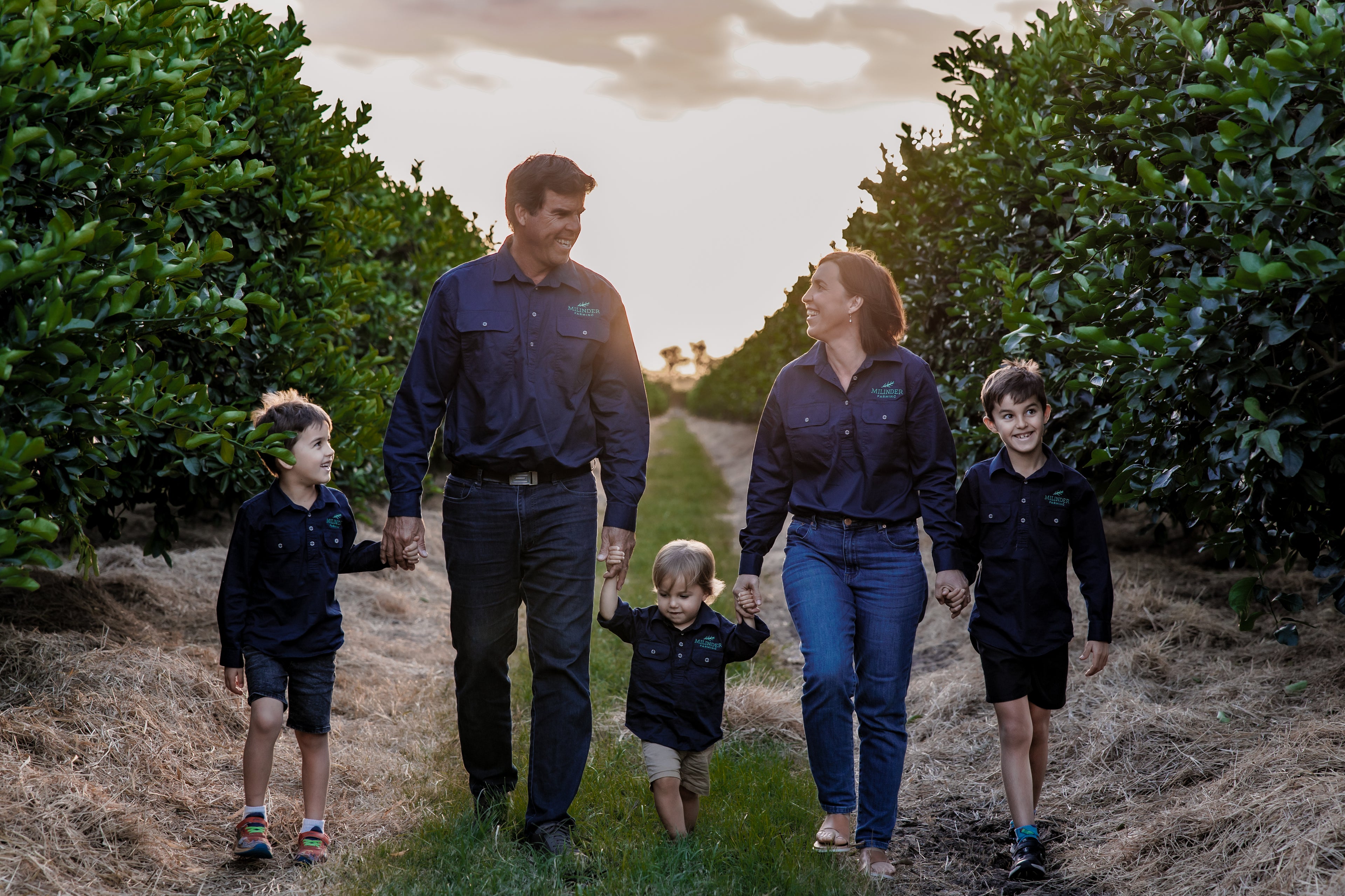 A family of five walking on their farm in Far North Queensland, Australia. The parents are holding hands, while the three children walk happily with them. The sun is setting and golden clouds are visible in the background.